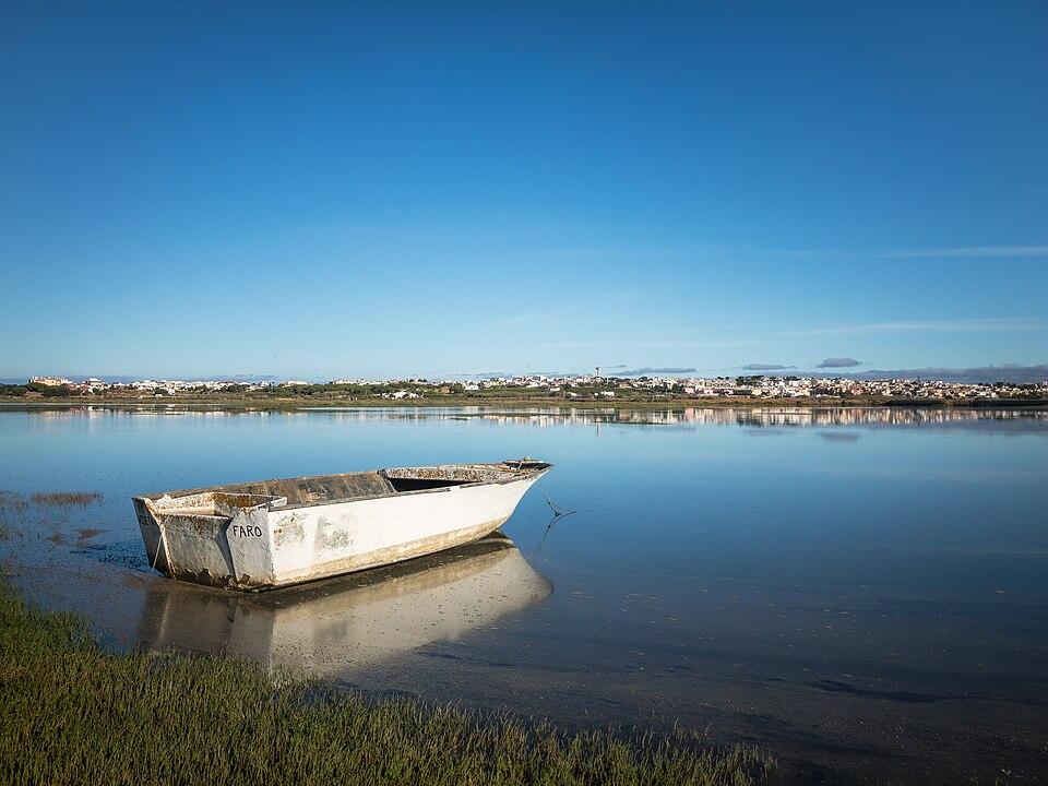 Pequeñas embarcaciones ancladas en la laguna de Ria Formosa, Faro, Algarve, Portugal, reflejándose en tranquilas aguas turquesas bajo un cielo azul claro.