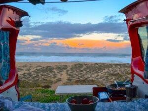 View from a red Ocean Camper van at sunset, beach, waves and a cooking station framed by the open doors.