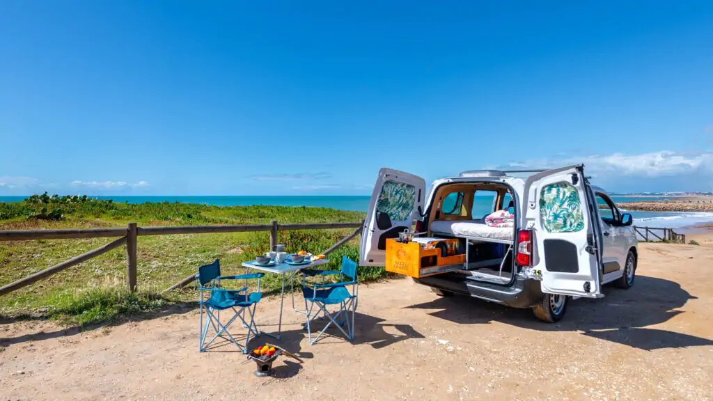 Indigo-Mini-Campervan mit geöffneter Seitentür, bezogenem Bett und sichtbarer Küchenzeile