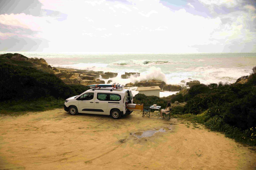 Surfařský obytný vůz Ocean Camper zaparkovaný na pobřeží Atlantiku v Portugalsku se surfovými prkny na střeše a cestujícím odpočívajícím u vln.