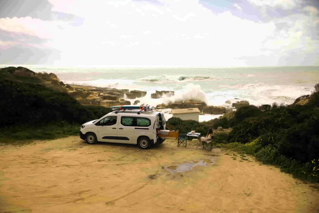 En surfcampervan från Ocean Camper som står parkerad vid Atlantkusten i Portugal, med surfbrädor på taket och en resenär som kopplar av vid vågorna.