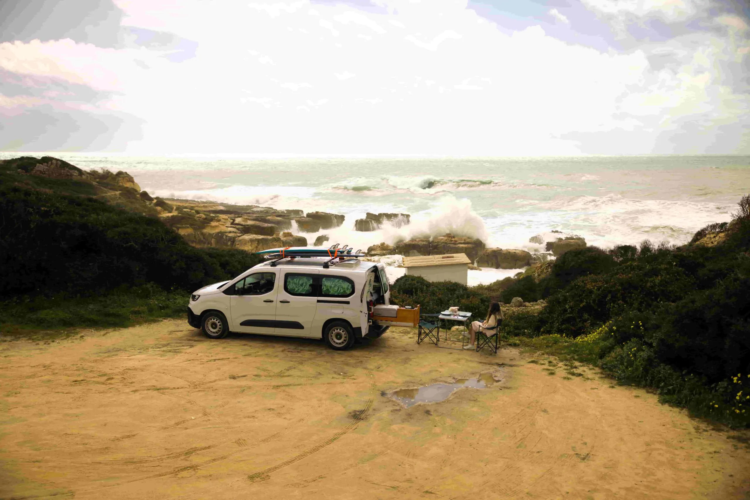 Una camper de surf de Ocean Camper aparcada en la costa atlántica de Portugal, con tablas de surf en el techo y un viajero relajándose junto a las olas.