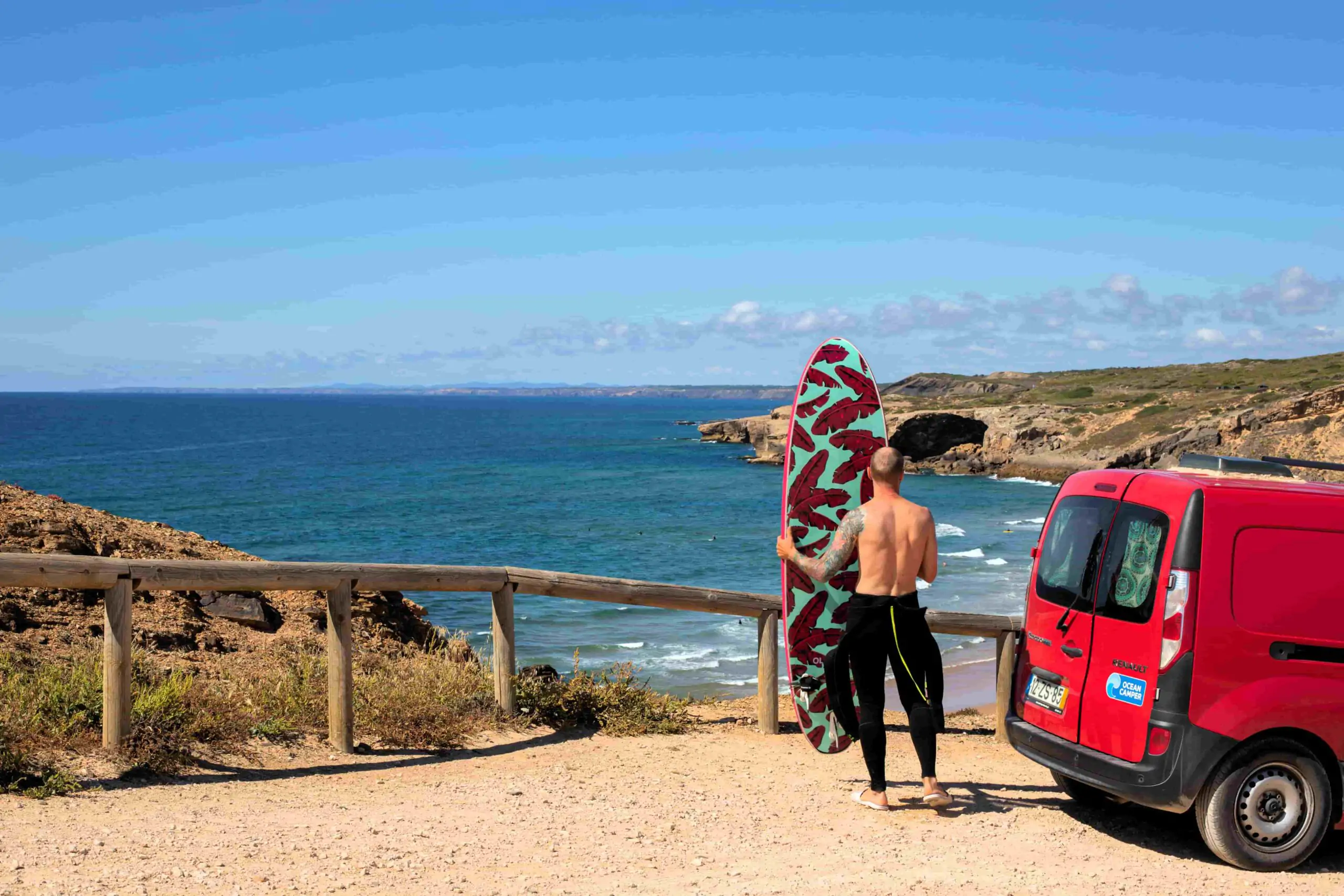 Un surfista con una tabla de surf roja junto a una furgoneta camper, con vistas a la costa atlántica de Portugal.
