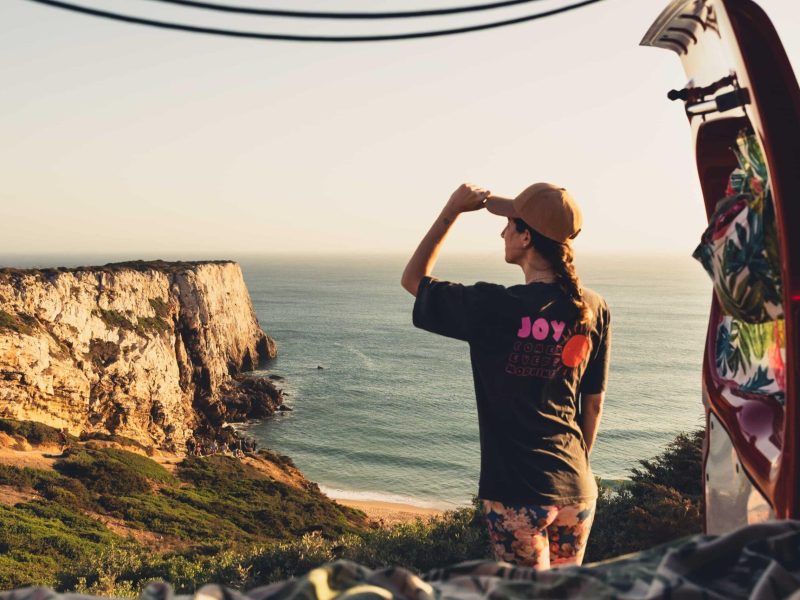 Woman standing at a campervan overlooking cliffs and the sea in Portugal during a coastal road trip.