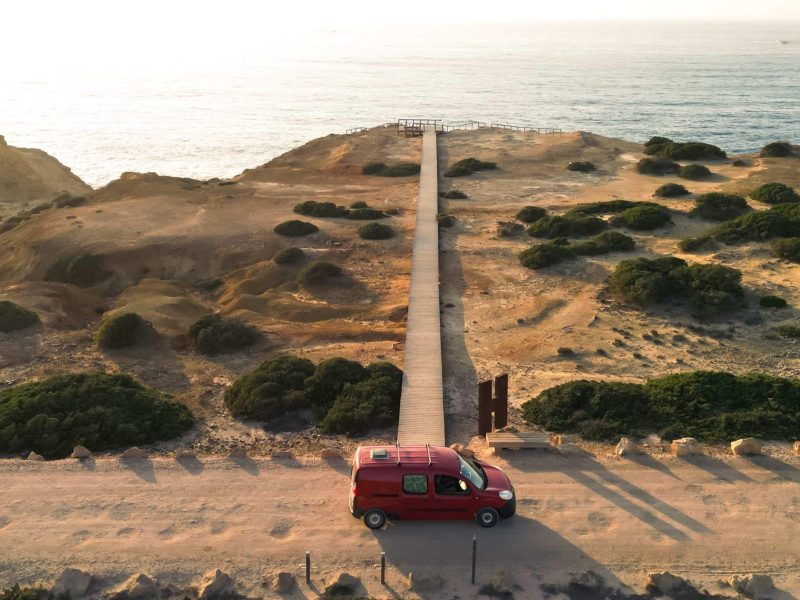 Red mini campervan parked by a coastal boardwalk overlooking cliffs and the ocean in Portugal.