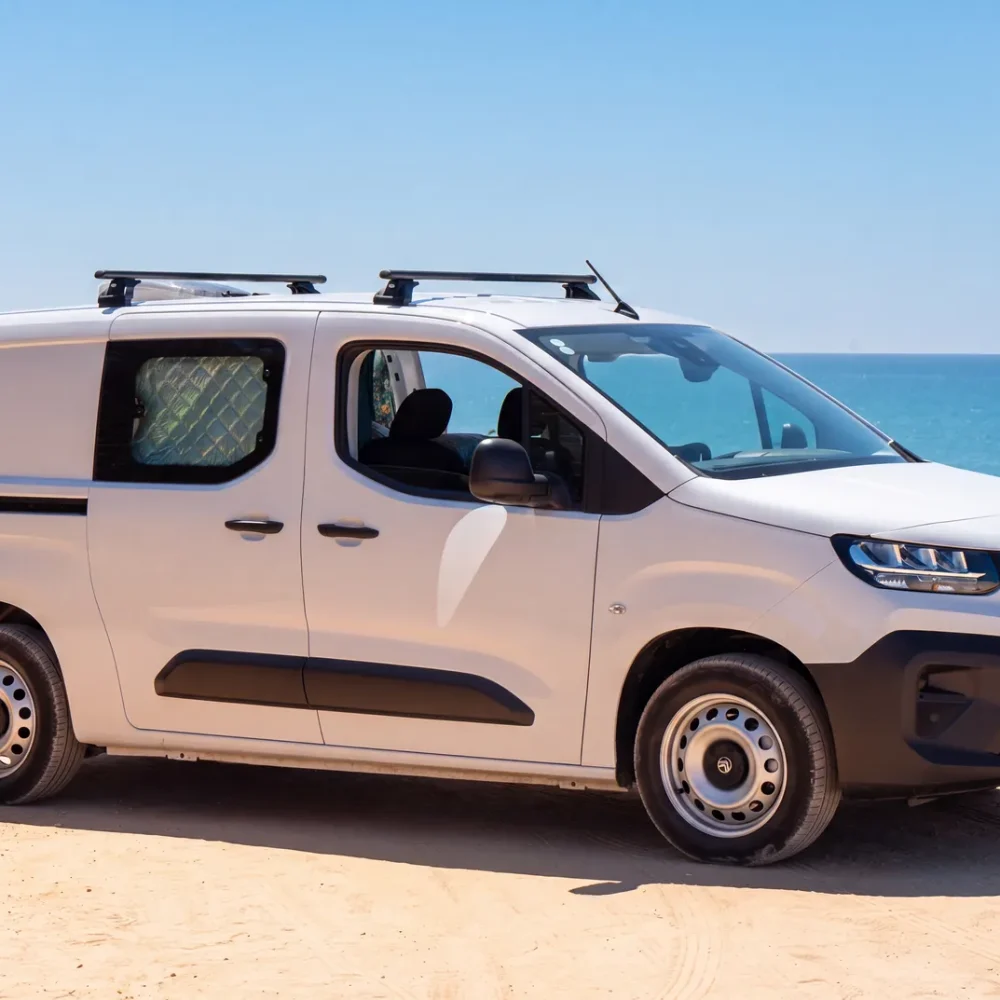 Bílý campervan Citroën Berlingo se střešními ližinami zaparkovaný na písku u moře